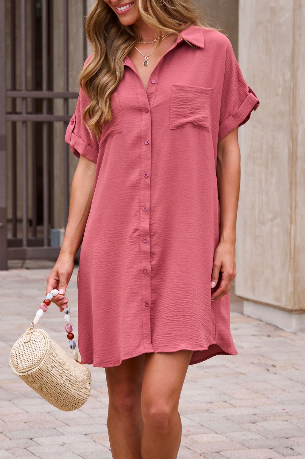 Woman in the Easy Button Up Shirt Dress - Terracotta holds a round straw handbag—perfect as a beach cover-up.