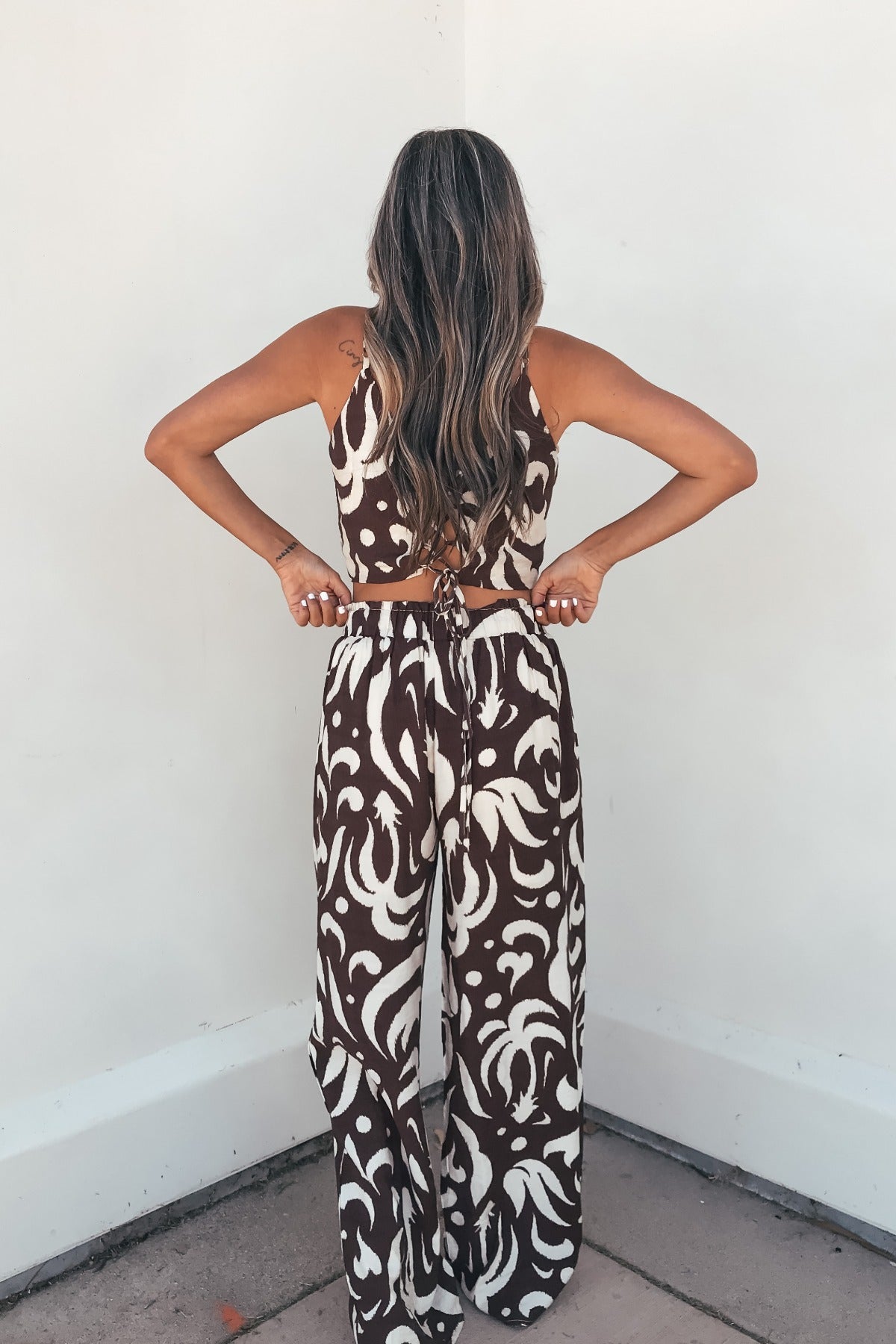 A woman with long wavy hair wears the Eda Print Cami Tank and Pants Set - Brown, perfect for vacation, facing a white wall.