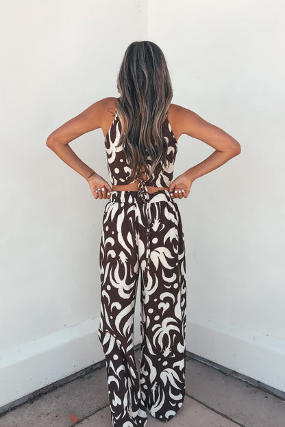 A woman with long wavy hair wears the Eda Print Cami Tank and Pants Set - Brown, perfect for vacation, facing a white wall.