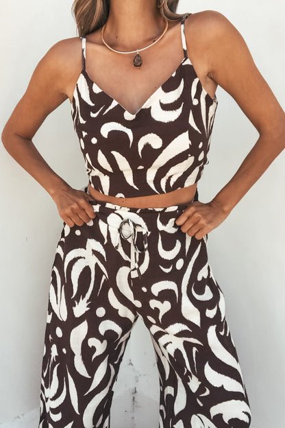 Woman in the Eda Print Cami Tank and Pants Set - Brown stands hands on hips against a white wall—a perfect vacation look.