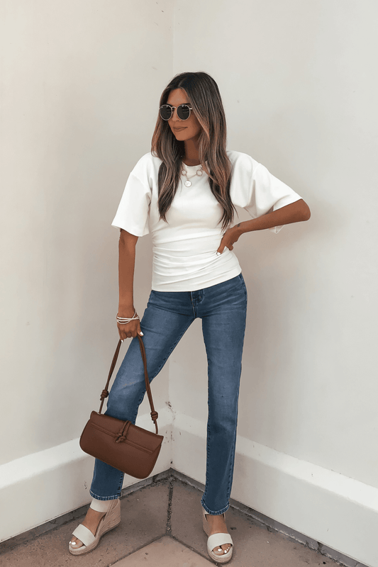 Woman in the Effortless Everyday Ruched Top - White, blue jeans, wedges, and brown bag stands against a white wall.