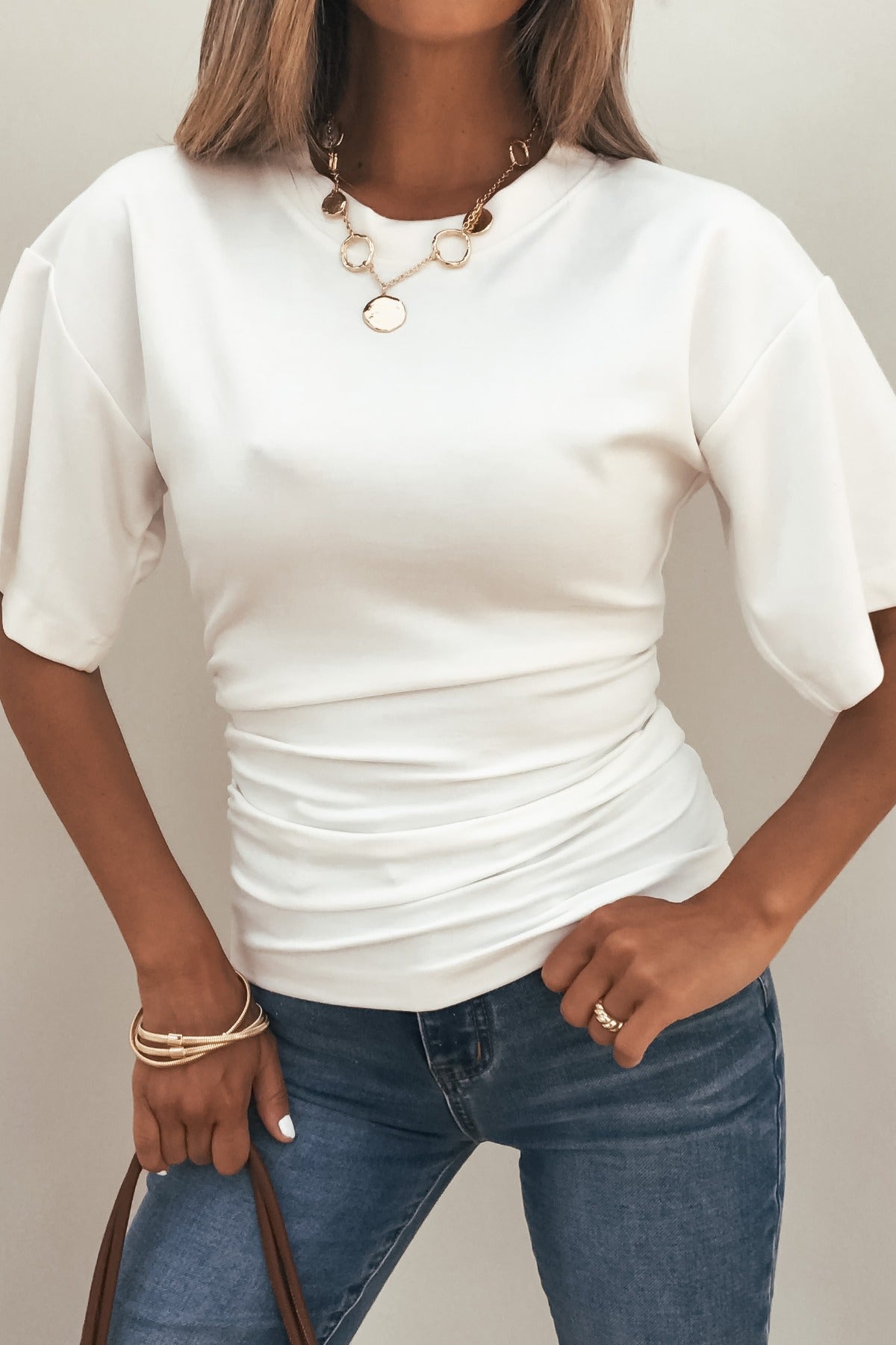 Woman wearing the Effortless Everyday Ruched Top - White, styled with blue jeans, gold jewelry, and a brown strap.