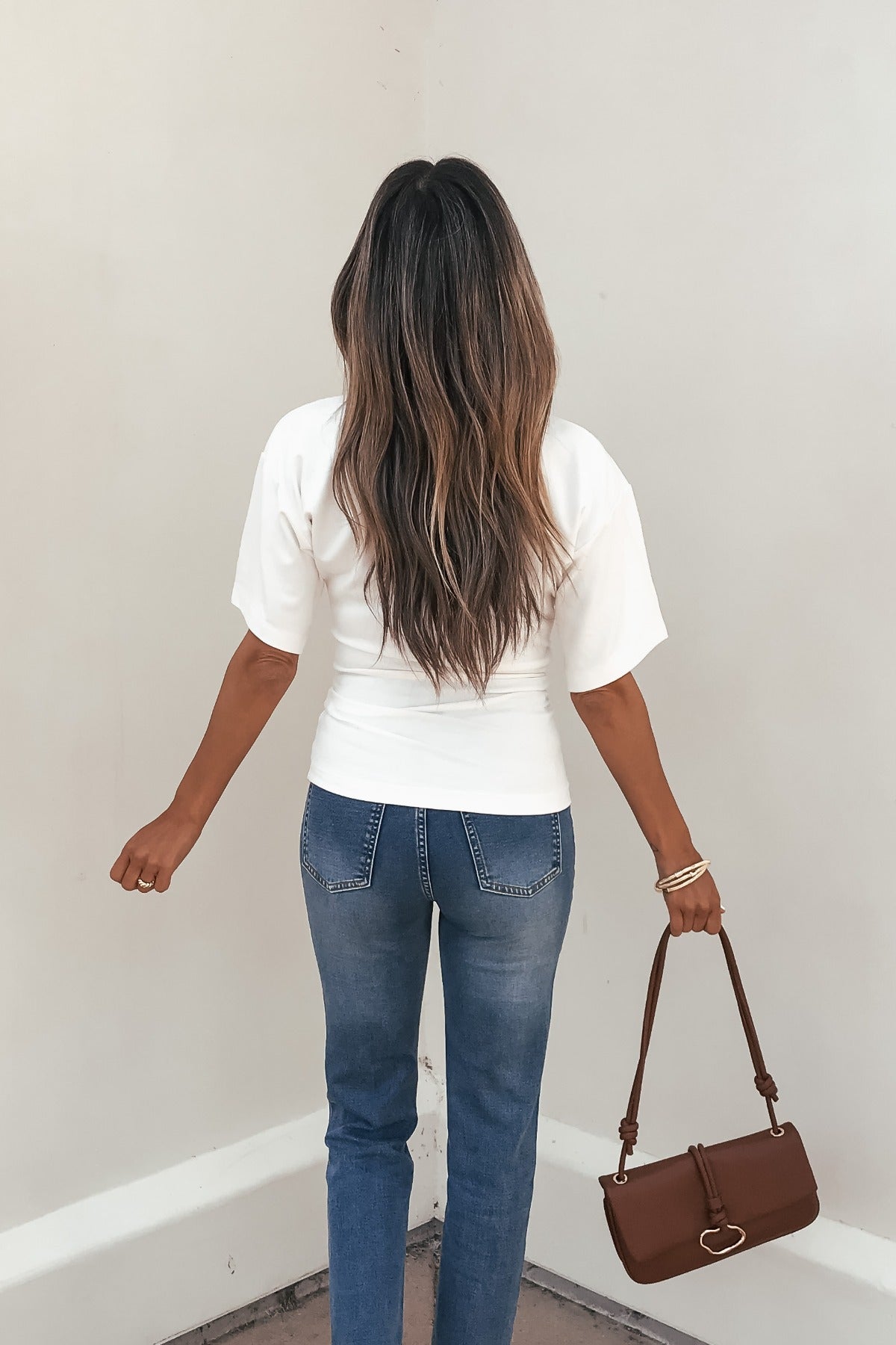 A woman with long wavy hair, in blue jeans and the Effortless Everyday Ruched Top - White, stands holding a brown handbag.