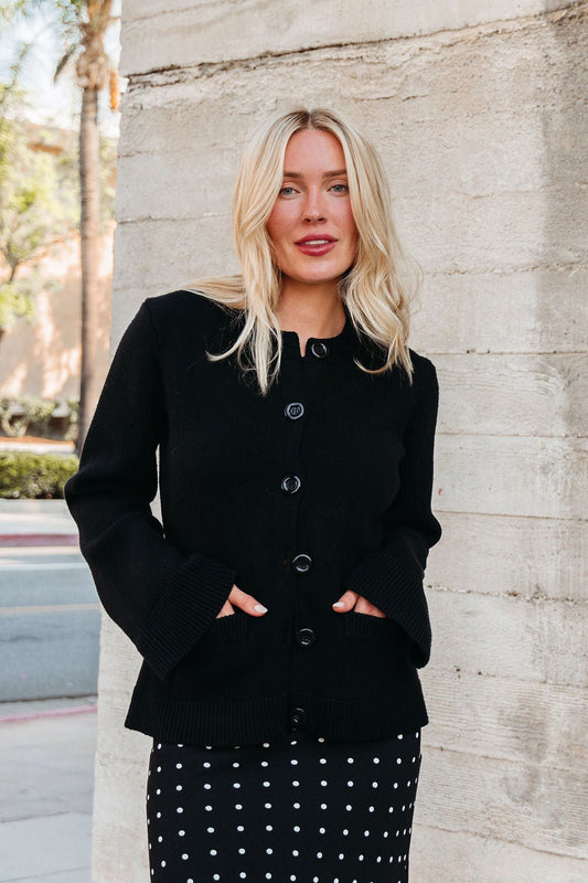 A woman with blonde hair wears the Elan Black Button Up Cardigan Jacket, standing outdoors against a stone wall, smiling.