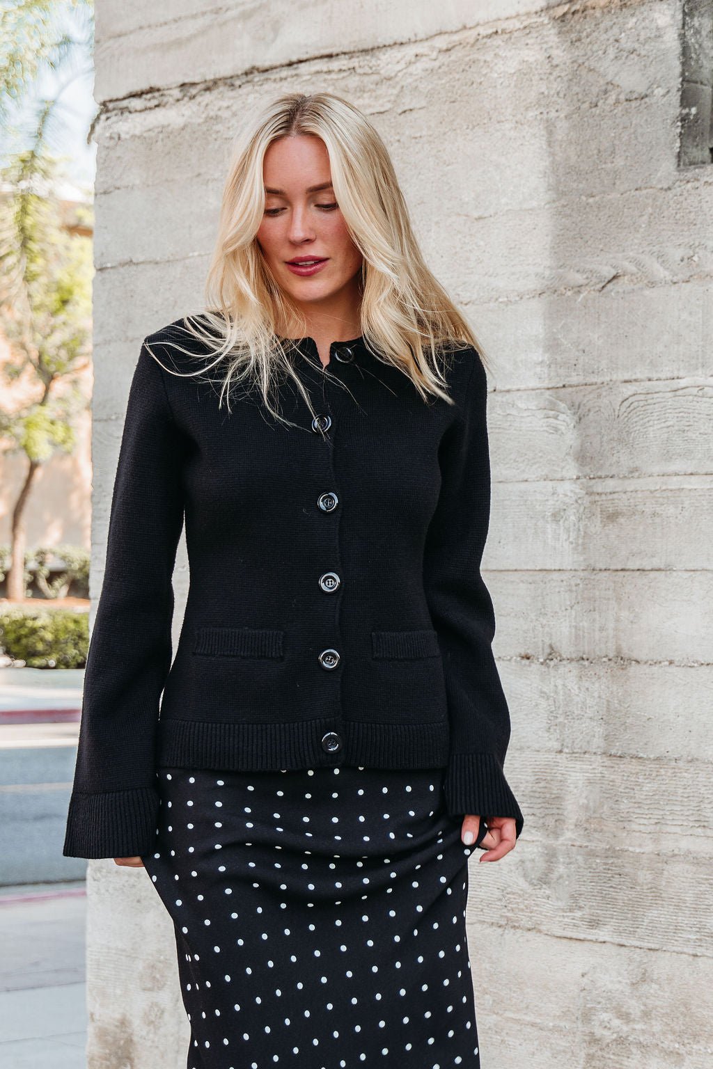A woman with long blonde hair wears the Elan Black Button Up Cardigan Jacket, standing by a concrete wall.