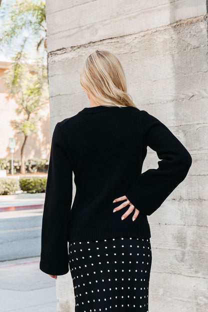 A woman with blonde hair stands outdoors, wearing the Elan Black Button Up Cardigan Jacket and a black skirt with white polka dots.