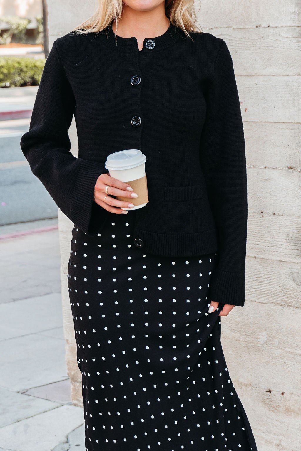Woman wears the Elan Black Button Up Cardigan Jacket with a polka dot skirt, holding coffee outdoors.