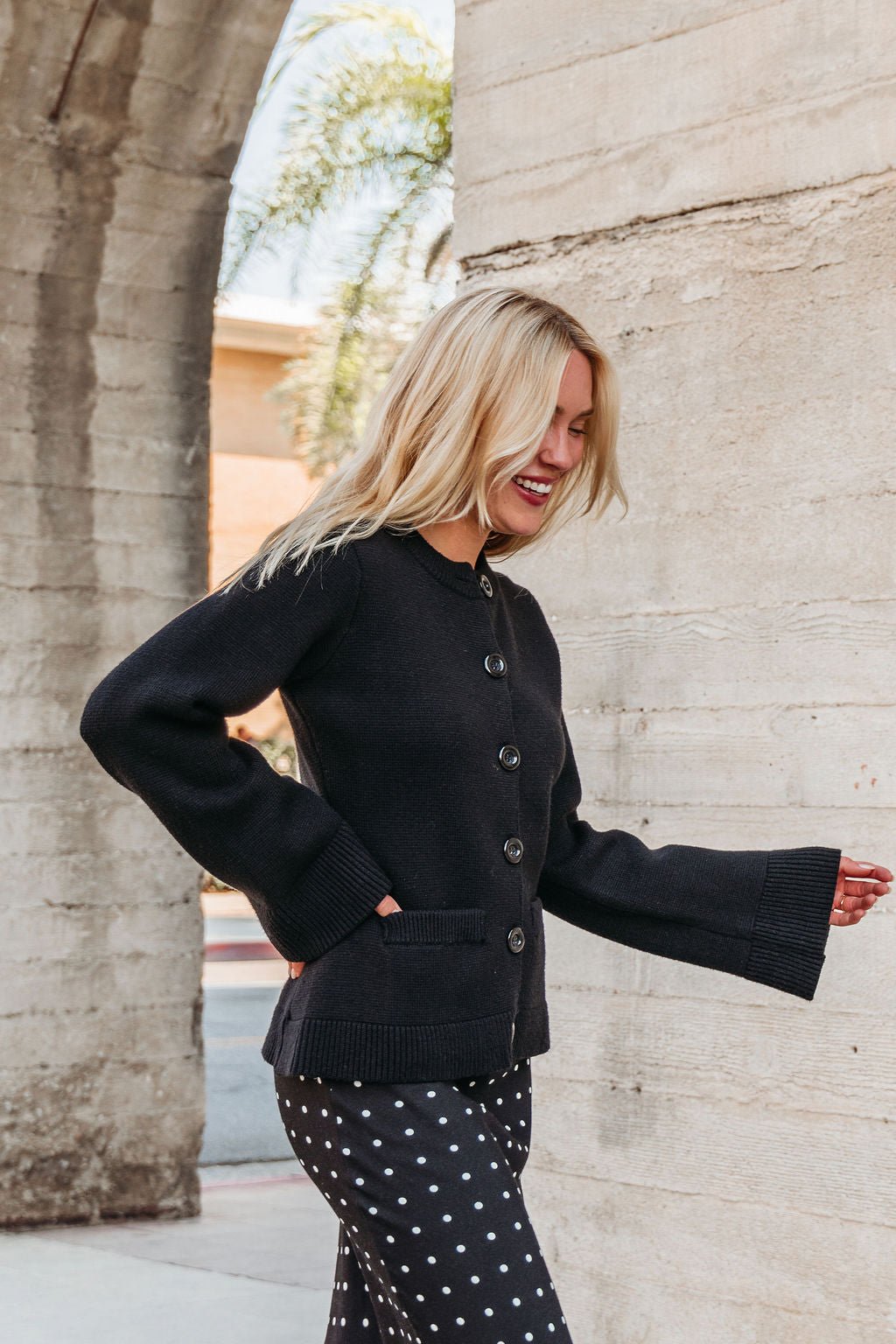 A woman with long blonde hair smiles outdoors, wearing the Elan Black Button Up Cardigan Jacket and black polka dot pants.