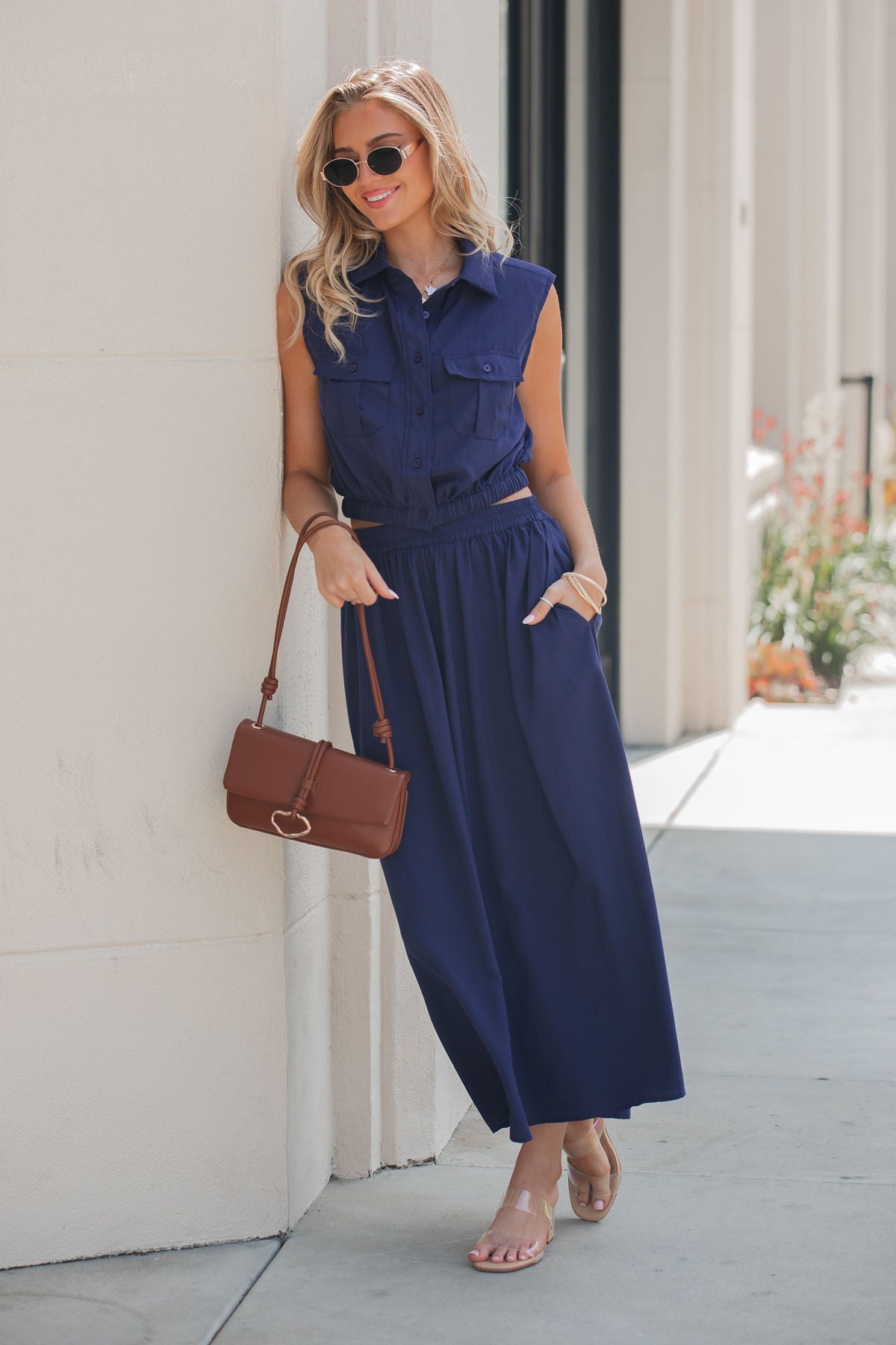Woman in an Elan Collared Button Down Top - Navy, sunglasses, and sandals, holding a brown purse outdoors against a light wall.
