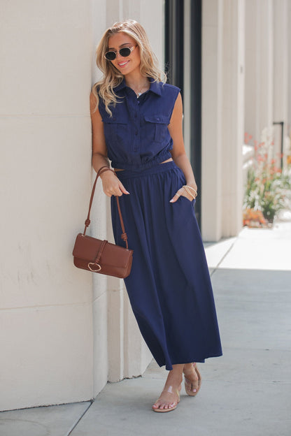 Woman in an Elan Collared Button Down Top - Navy, sunglasses, and sandals, holding a brown purse outdoors against a light wall.