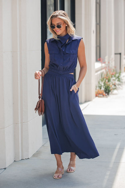 Woman in an Elan Collared Button Down Top - Navy, sunglasses, and sandals holds a brown bag, walking by a light building.