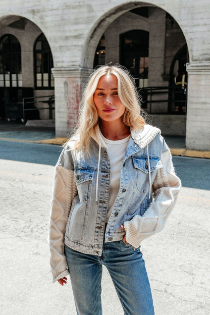 A woman with long blonde hair wears the Elan Denim Hooded Sweater Jacket outdoors by an arched building.
