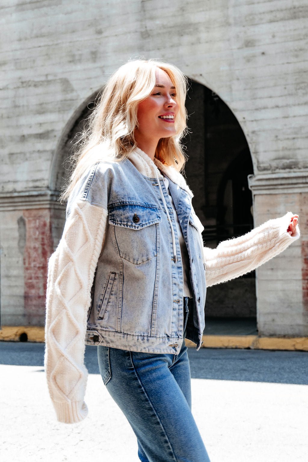 A smiling woman in the Elan Denim Hooded Sweater Jacket walks outdoors by a concrete wall with arched openings.