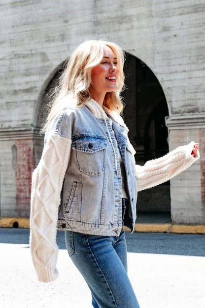A smiling woman in the Elan Denim Hooded Sweater Jacket walks outdoors by a concrete wall with arched openings.