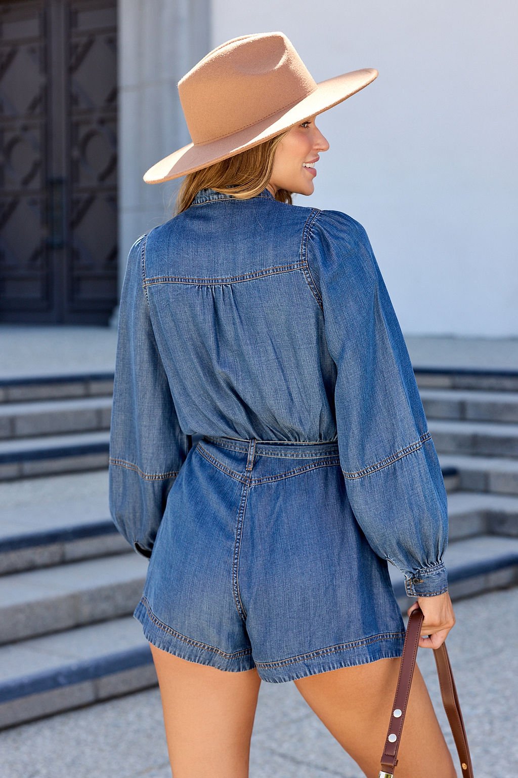 Woman wears the Elan Denim Long Sleeve Belted Romper, tan wide-brim hat, and holds a brown bag near outdoor stone steps.