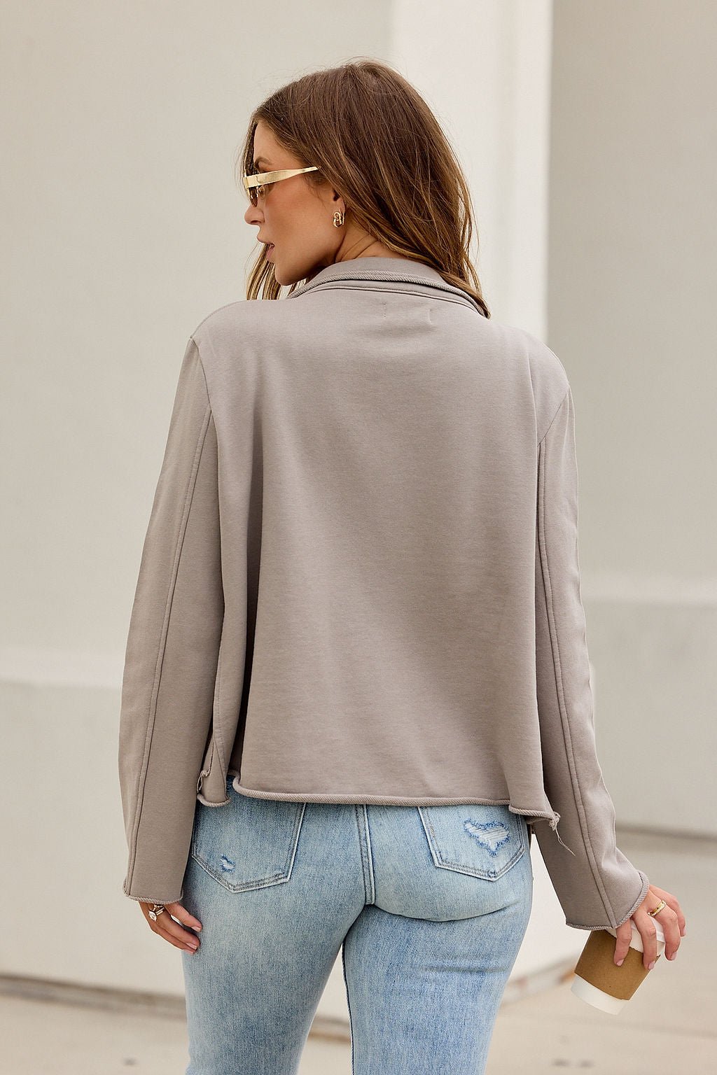 Woman with light brown hair in sunglasses and the ELAN Double Button Jacket - Taupe, ripped jeans, holding coffee, seen from behind.