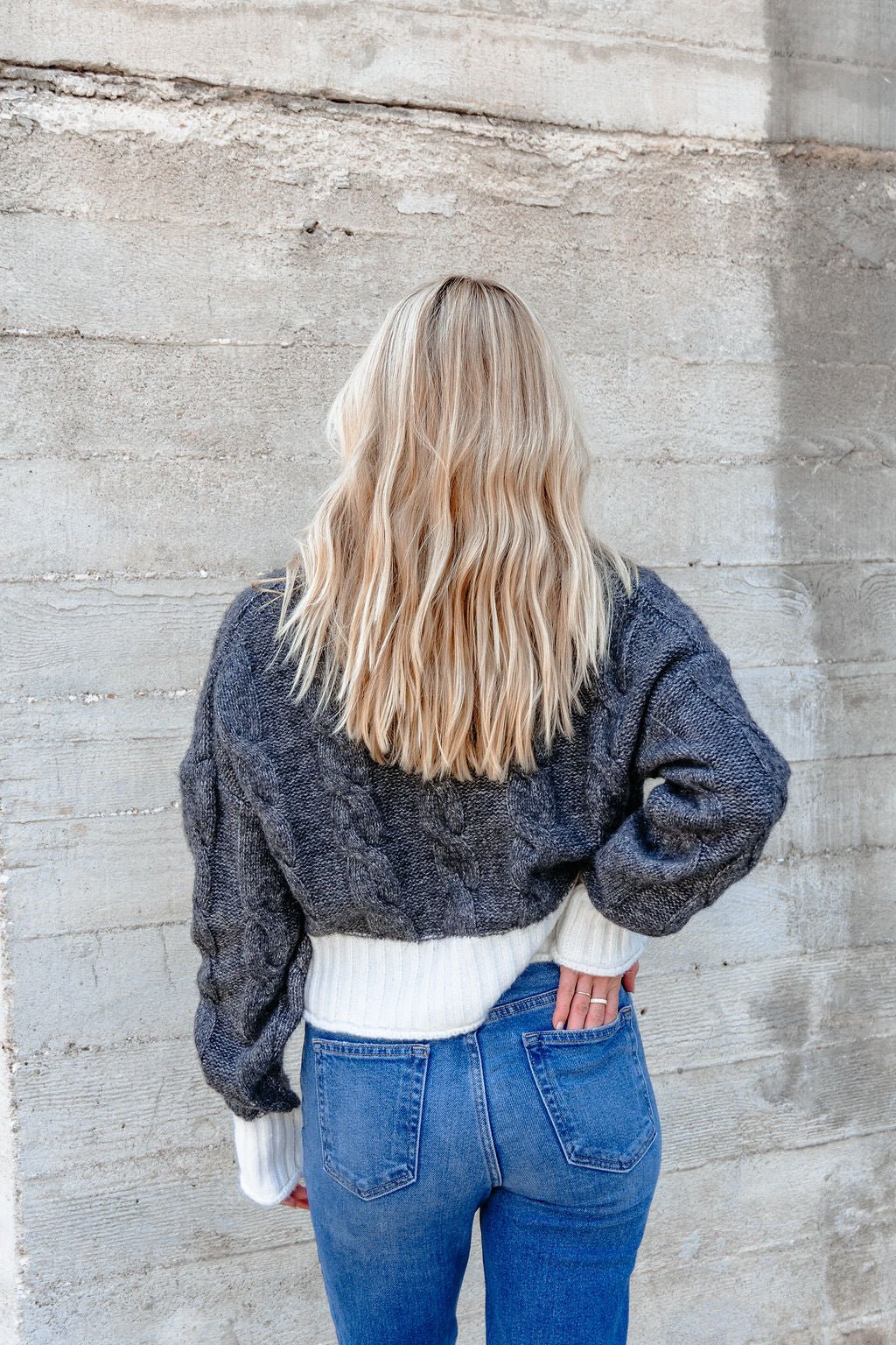 A woman wears the Elan Grey and White Cable Knit Sweater, standing by a concrete wall with her hand in her pocket.