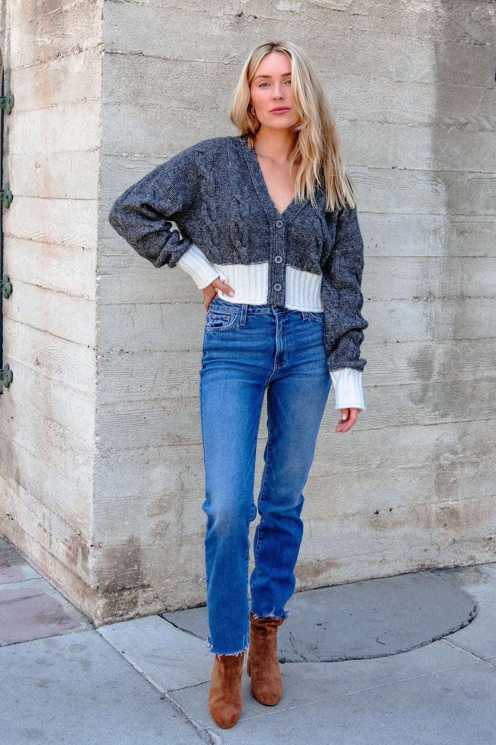 A woman wears the Elan Grey and White Cable Knit Sweater, blue jeans, and brown boots by a textured concrete outdoor wall.