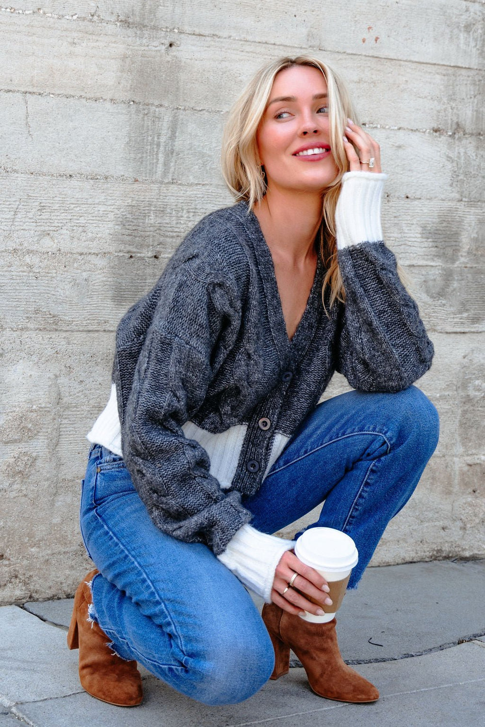 A woman in an Elan Grey and White Cable Knit Sweater squats by a wall, smiling with a coffee cup.