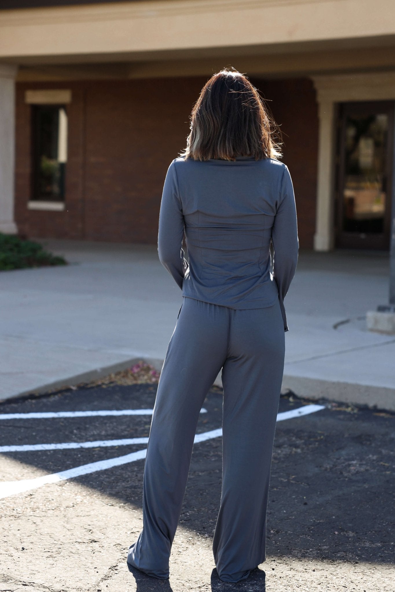 A woman faces away outdoors in a parking lot, wearing Elan Grey Flowy Wide Leg Pants - DOORBUSTER and a gray long-sleeve top.