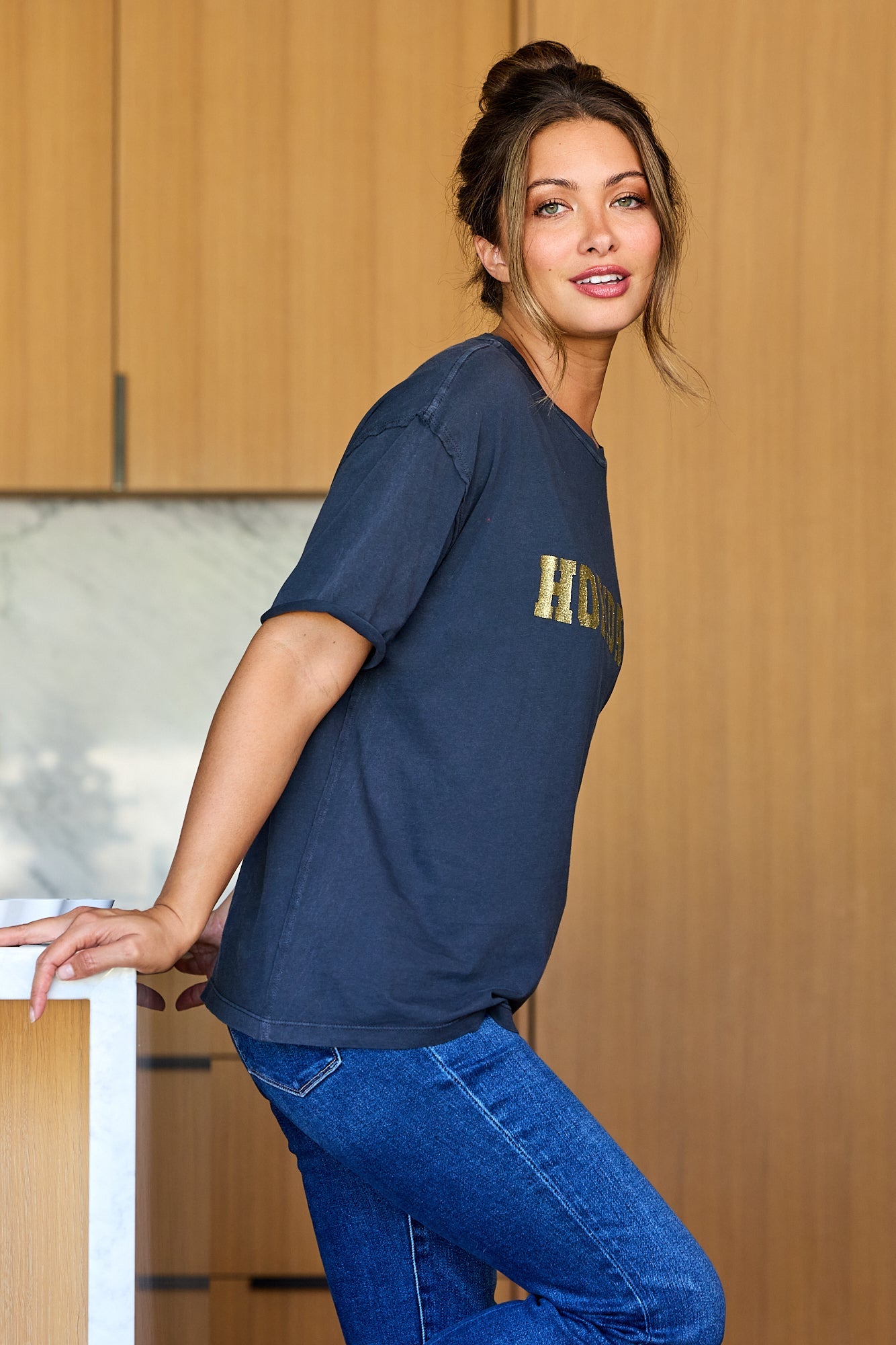 A woman wears an Elan Grey Holiday Graphic Tee, blue jeans, and a bun, smiling at the camera while leaning on a kitchen counter.