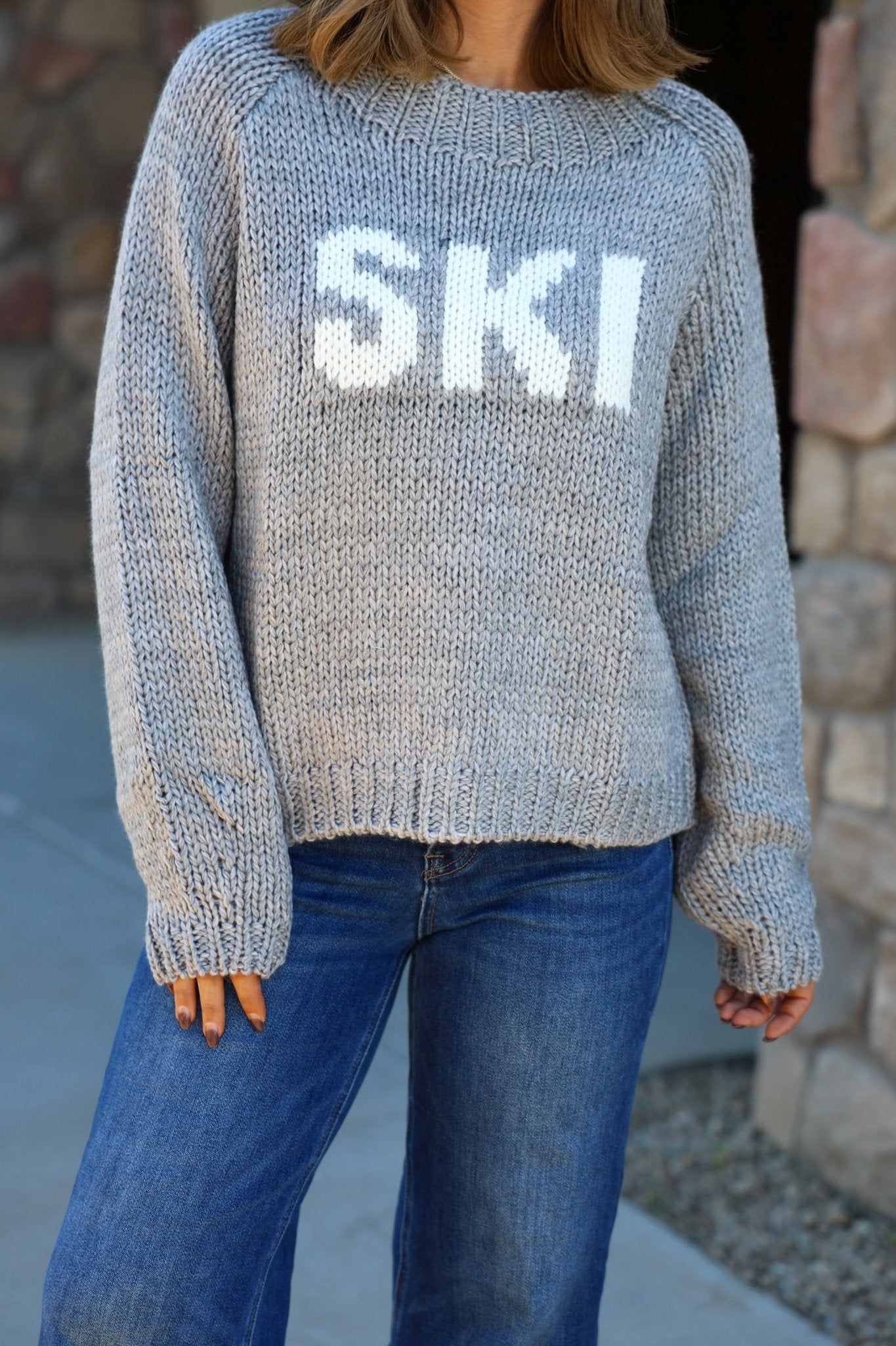 Someone in the Elan Grey Ski Ribbed Sweater and blue jeans stands by a stone wall.