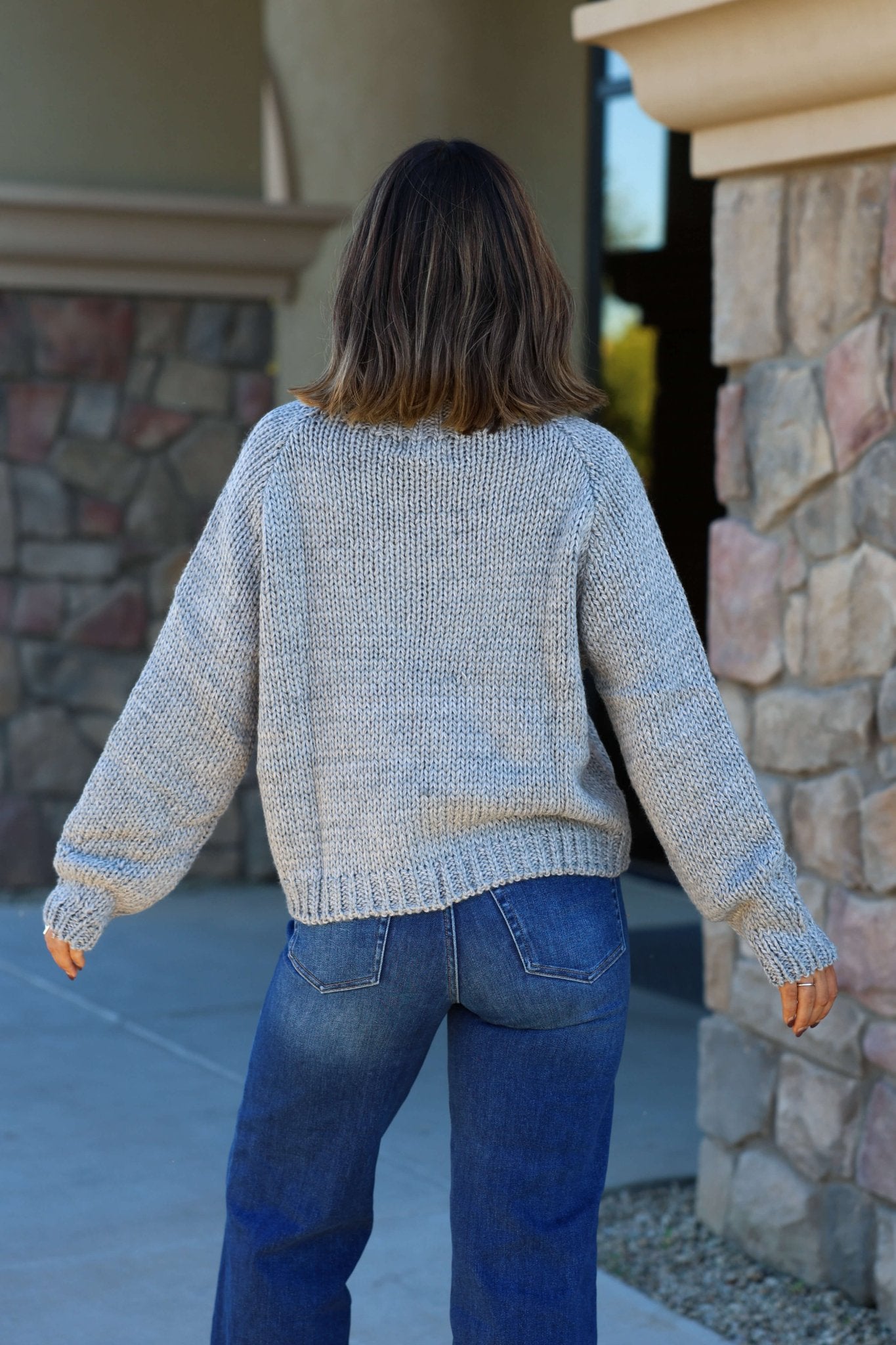 Wearing an Elan Grey Ski Ribbed Sweater and blue jeans, a person stands outside by a stone building, facing away.