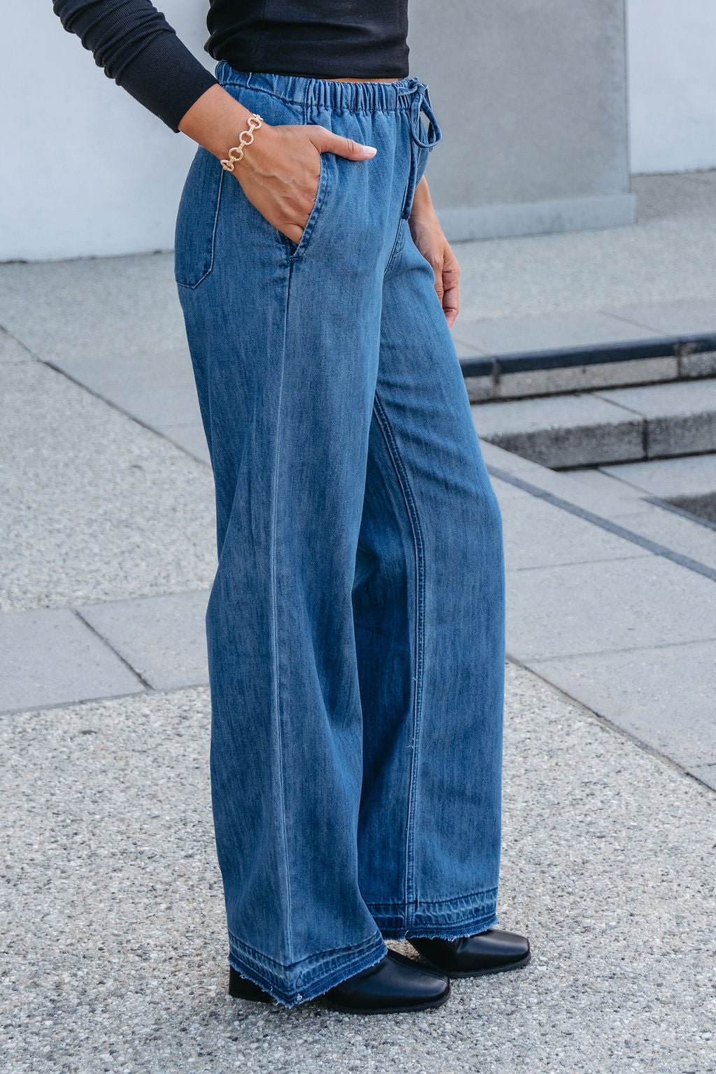 Someone wearing Elan Medium Wash Wide Leg Pants, a black top, and black shoes stands outdoors with one hand in their pocket.