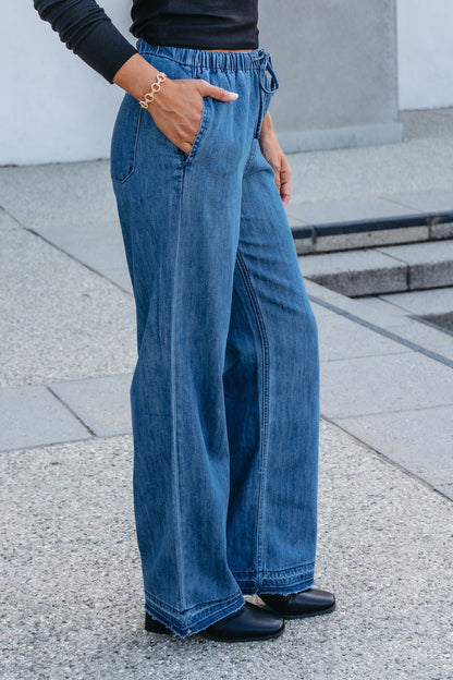 Someone wearing Elan Medium Wash Wide Leg Pants, a black top, and black shoes stands outdoors with one hand in their pocket.