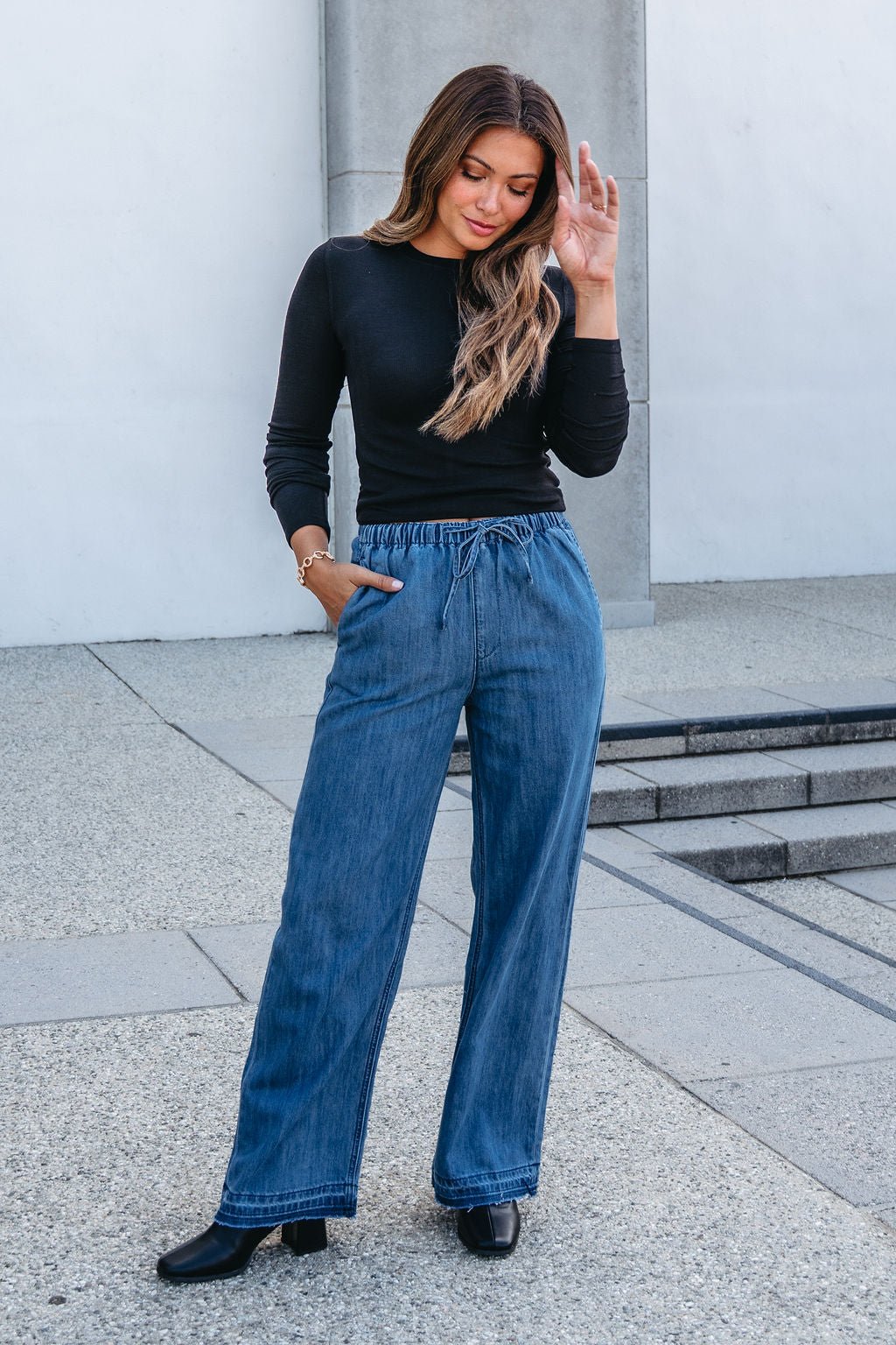 Woman wearing a black long-sleeve top, Elan Medium Wash Wide Leg Pants, and black boots, standing outdoors on concrete.