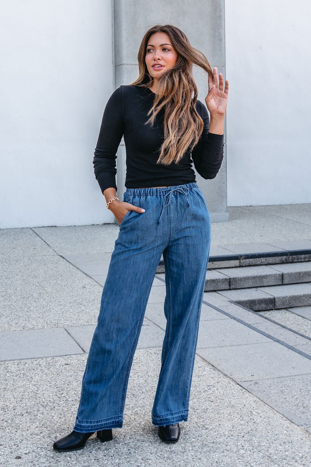 Woman wearing Elan Medium Wash Wide Leg Pants, a black long-sleeve top, and boots outdoors on a paved walkway.