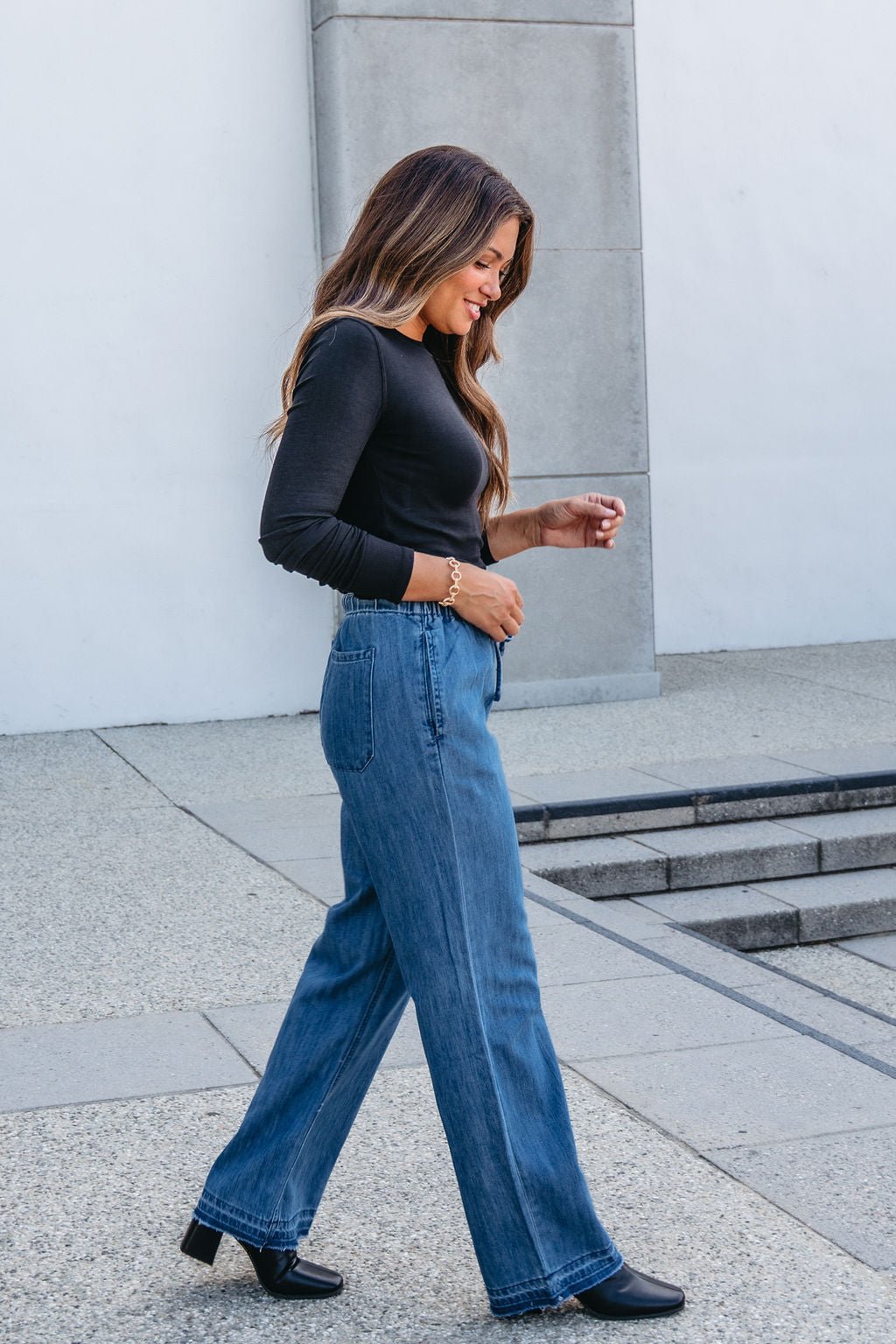 Smiling woman with long hair in a black top and Elan Medium Wash Wide Leg Pants walks outdoors.