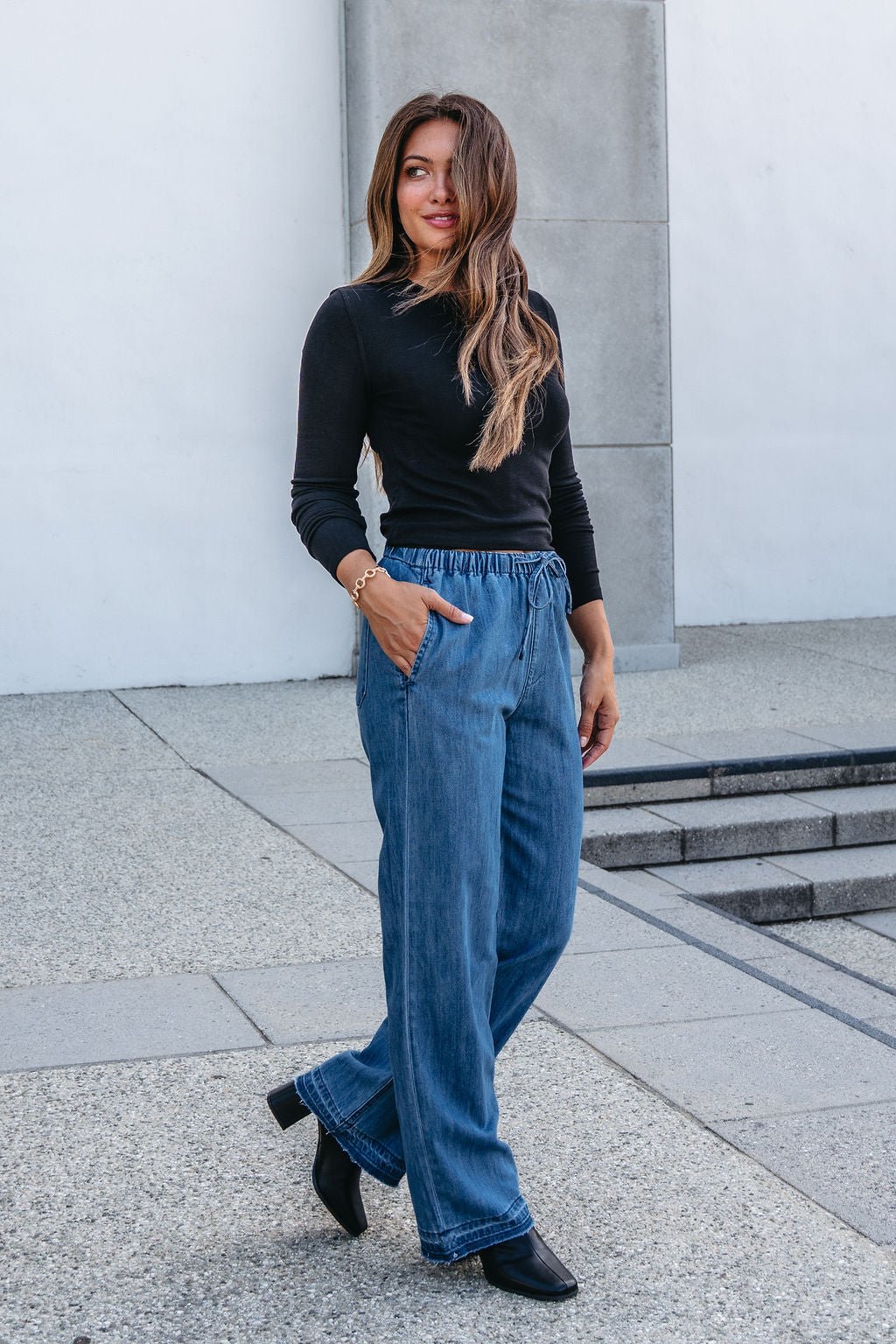 A woman in Elan Medium Wash Wide Leg Pants and a black long-sleeve top stands outside, smiling with a hand in her pocket.