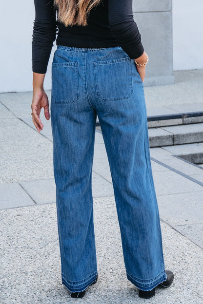 A woman wearing Elan Medium Wash Wide Leg Pants and a black top stands outdoors on concrete, facing away from the camera.