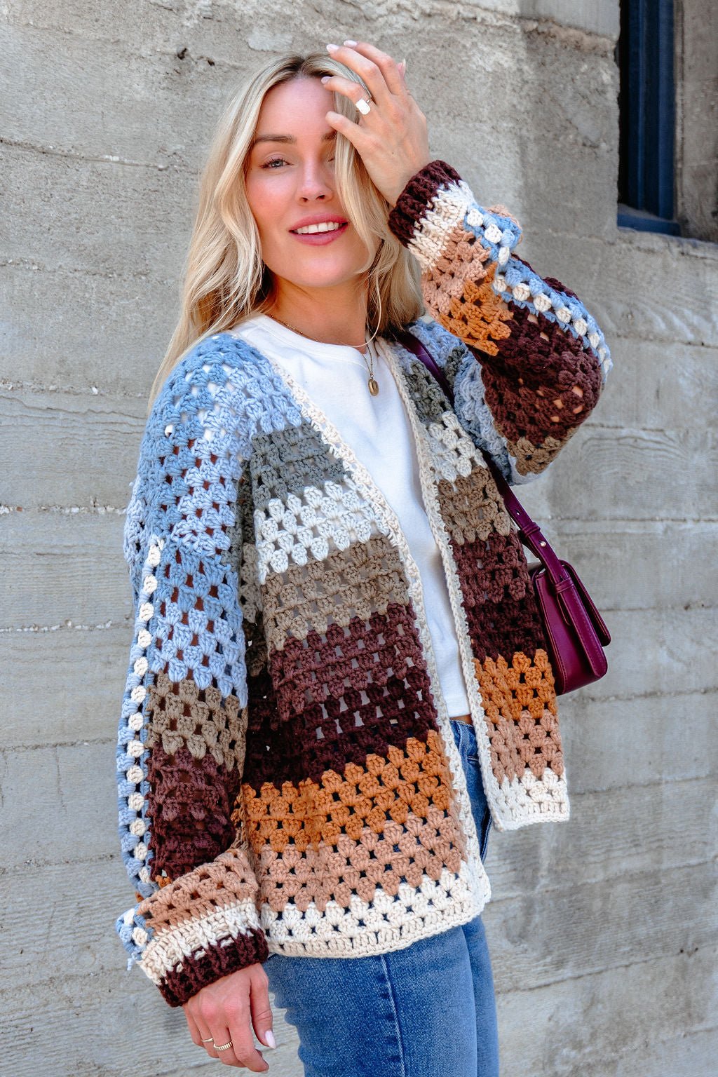 Woman smiling in the Elan Multi Striped Crochet Open Cardigan, white shirt, and jeans, holding a purple shoulder bag.