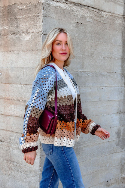 Woman with blonde hair wears the Elan Multi Striped Crochet Open Cardigan, blue jeans, and a maroon purse by a concrete wall.