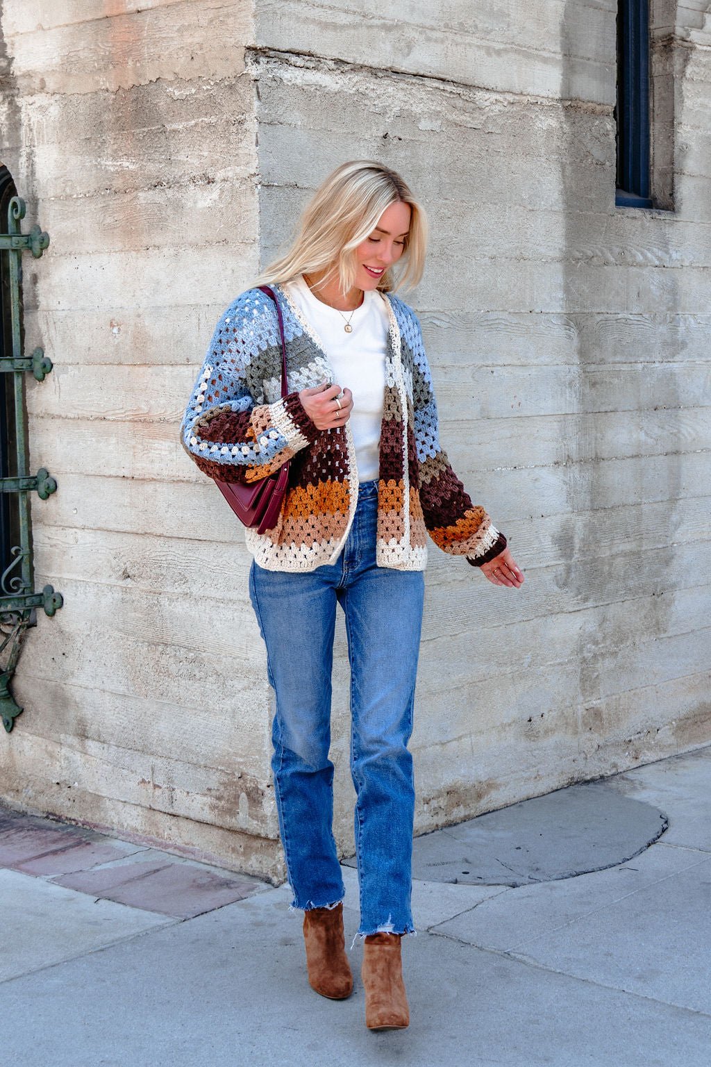 A woman wears the Elan Multi Striped Crochet Open Cardigan, blue jeans, and brown boots while walking by a concrete wall.