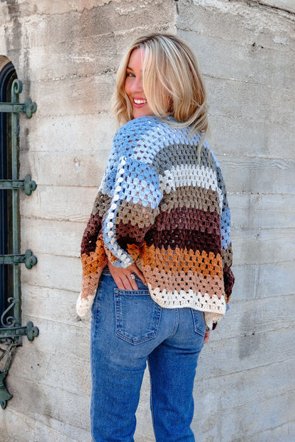 Smiling, a blonde woman wears the Elan Multi Striped Crochet Open Cardigan—FINAL SALE—with blue jeans by a concrete wall.