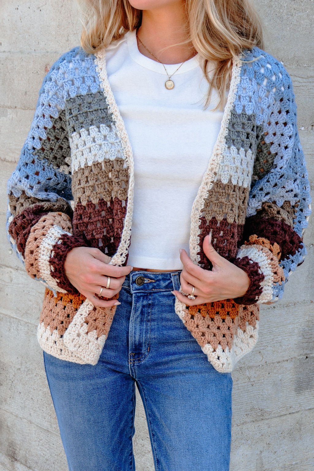 A woman models the Elan Multi Striped Crochet Open Cardigan over a white tee and blue jeans by a concrete wall.