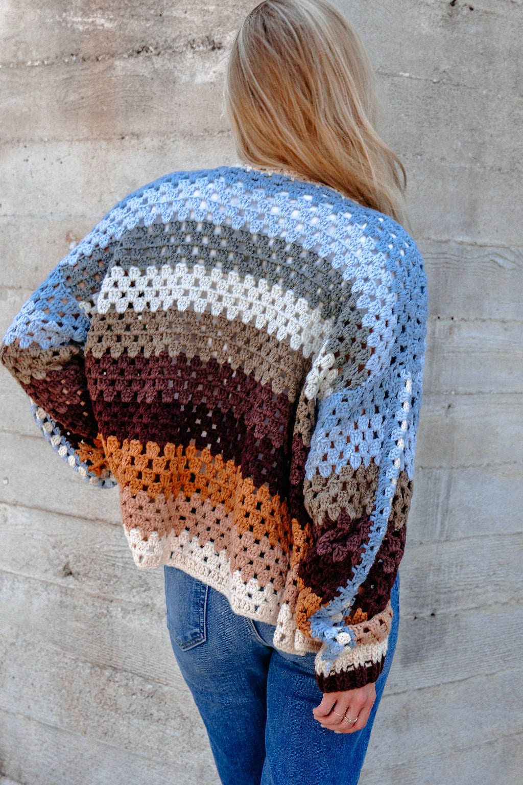 A woman with long blonde hair wears the Elan Multi Striped Crochet Open Cardigan, paired with blue jeans by a concrete wall.