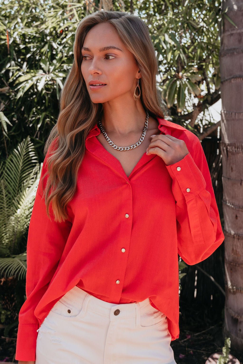 A woman in an Elan Red Button Down Collared Shirt and white pants stands outdoors among greenery and a palm tree.