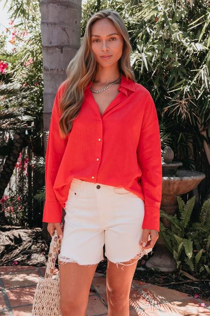 A woman in an Elan Red Button Down Collared Shirt and white denim shorts stands outside, holding a woven bag.
