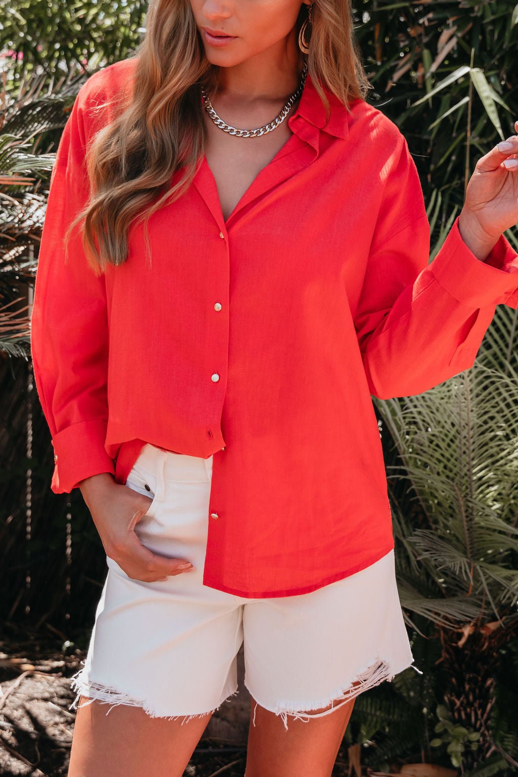 Woman in an Elan Red Button Down Collared Shirt and white frayed shorts, standing outdoors by green plants.