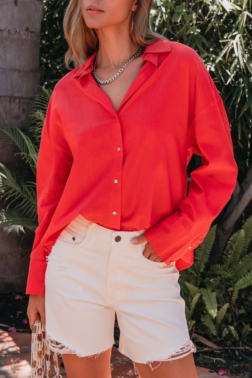 Woman wearing an Elan Red Button Down Collared Shirt and white frayed shorts, standing outdoors by green plants.