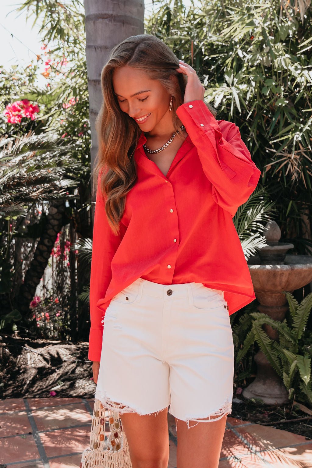 A woman wearing an Elan Red Button Down Collared Shirt and white shorts smiles outdoors with greenery in the background.