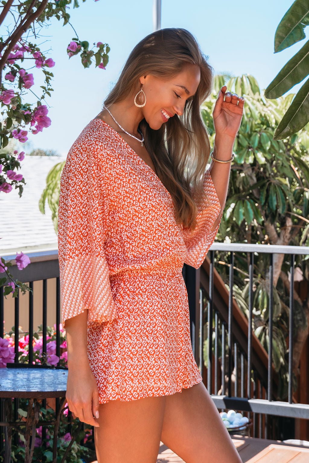 A woman smiles in the Elan Red Print Kimono Romper by tropical plants and flowers on a sunny day.