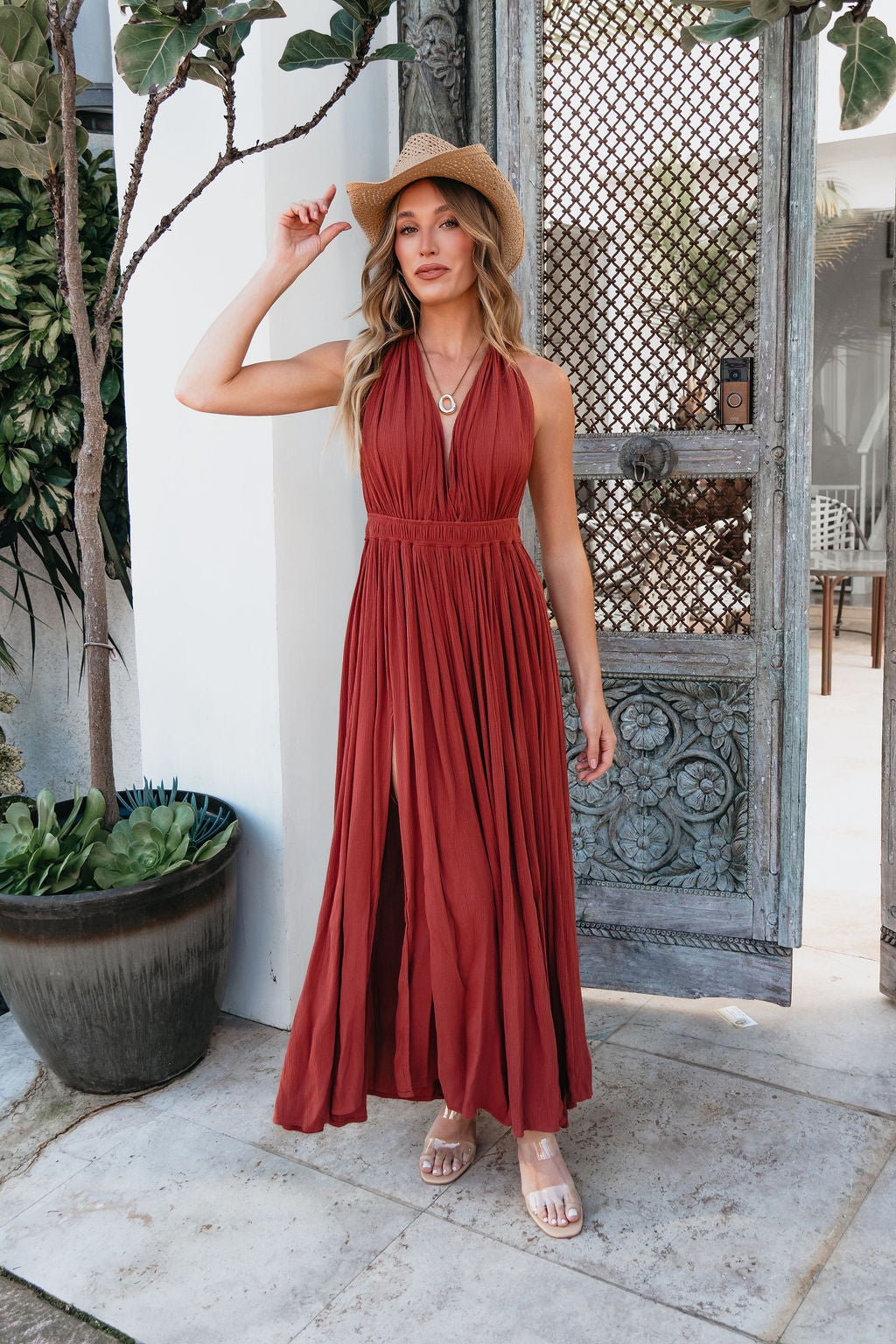 Woman in the ELAN Rust Slit Halter Maxi Dress and straw hat stands before an ornate door, holding her hat’s brim.