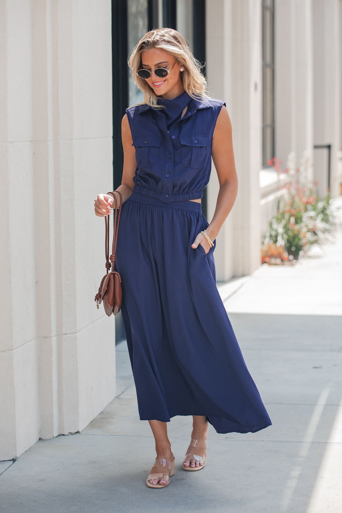 Woman in a navy Elan Smocked Midi Skirt, holding a brown bag, wearing sunglasses and sandals on a sunny sidewalk.
