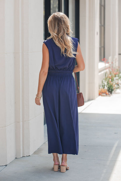 Woman with wavy blonde hair wears the Elan Smocked Midi Skirt in navy and pink sandals, carrying a brown purse on a sunny sidewalk.