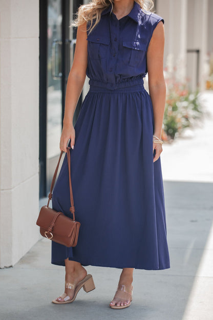 Woman wearing the Elan Smocked Midi Skirt in navy, a brown shoulder bag, and clear-heeled sandals on a sidewalk.